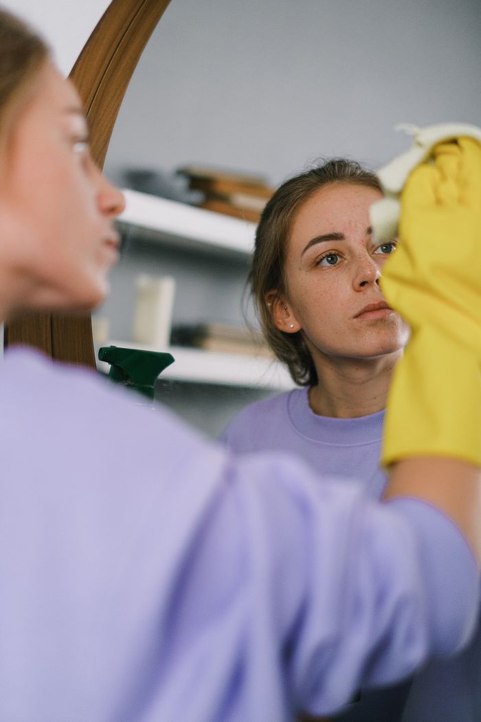 services-06 Crop female in purple sweatshirt and latex gloves washing mirror in light apartment in daytime